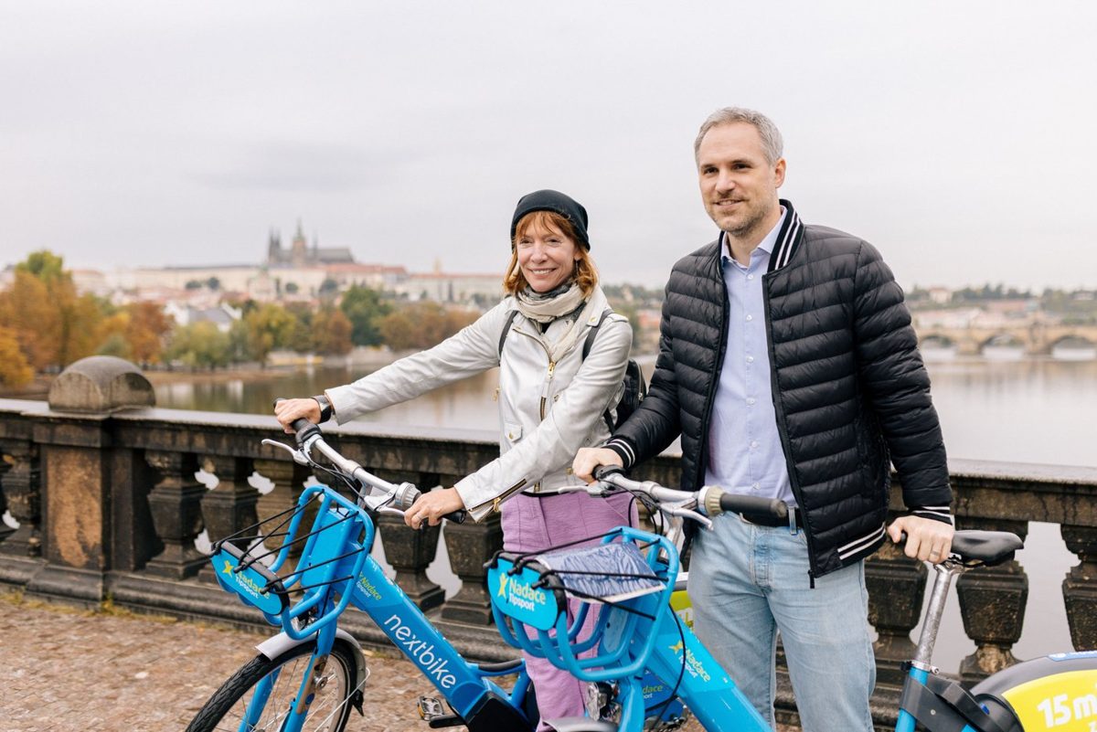 Dva lidé s modrými koly nextbike na nábřeží v Praze, v pozadí Pražský hrad